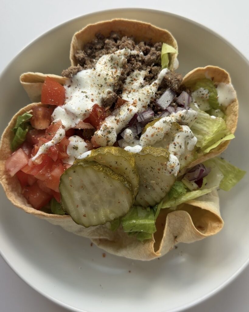 Close up of a burger-inspired meal featuring sliced pickles, chopped onions, tomatoes, lettuce, and seasoned toppings in a taco shell.