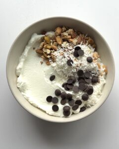 Bowl of yogurt topped with shredded coconut, chocolate chips, honey, cinnamon, nuts, and vanilla.