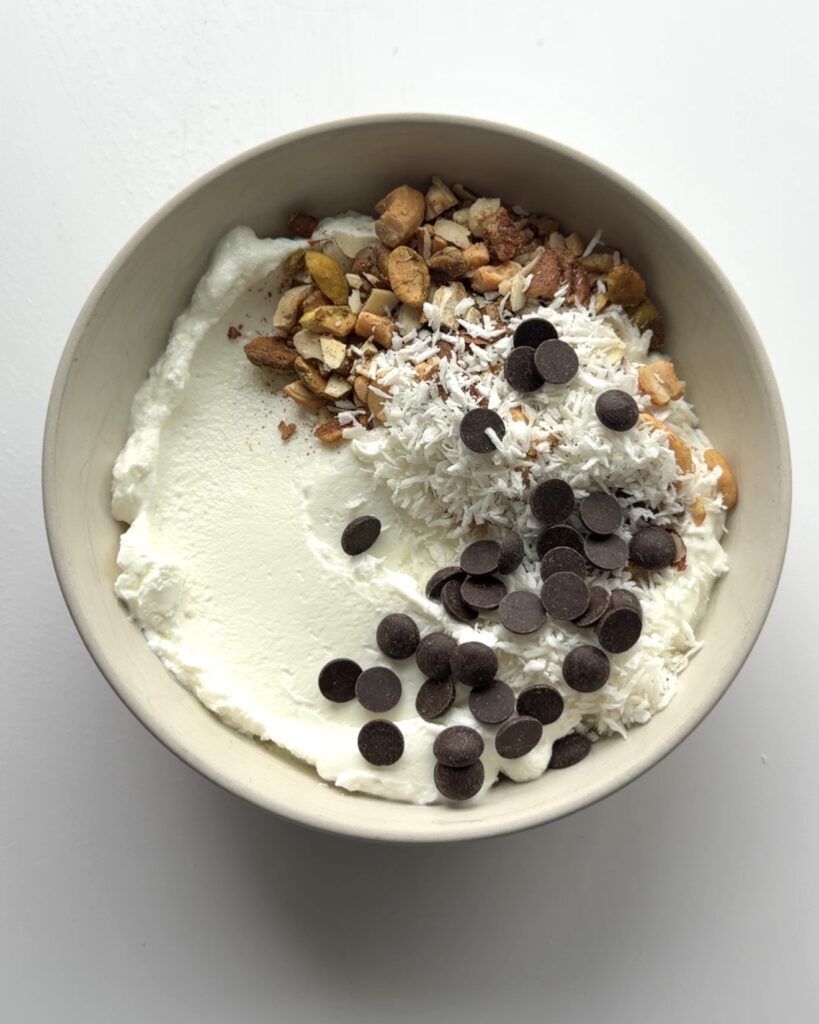 Bowl of yogurt topped with shredded coconut, chocolate chips, honey, cinnamon, nuts, and vanilla.