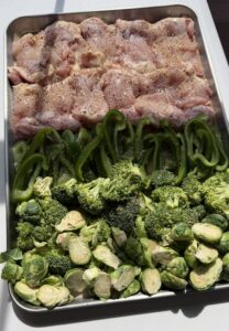 A sheet pan of raw chicken thighs with green peppers, broccoli, and Brussels sprouts.