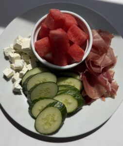 A plate with watermelon cubes, feta cheese, prosciutto, and sliced cucumber.