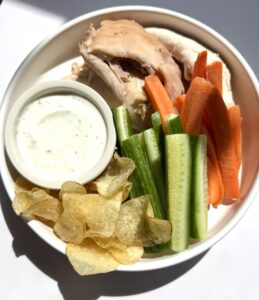 A plate with chicken, carrots, cucumber, chips, and dip.